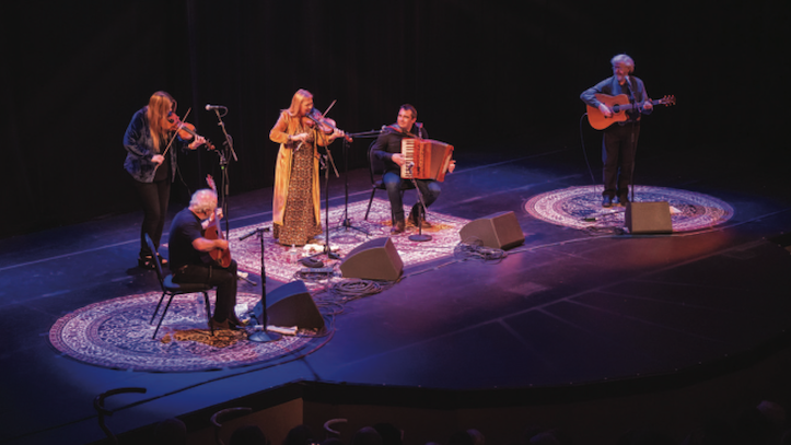Acoustic band performing live on stage on circular patterned rugs under blue stage spotlights.